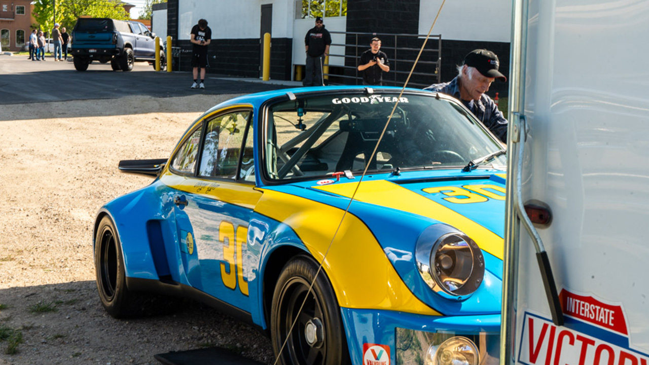 A blue and yellow vintage Porsche race car being unloaded from its trailer