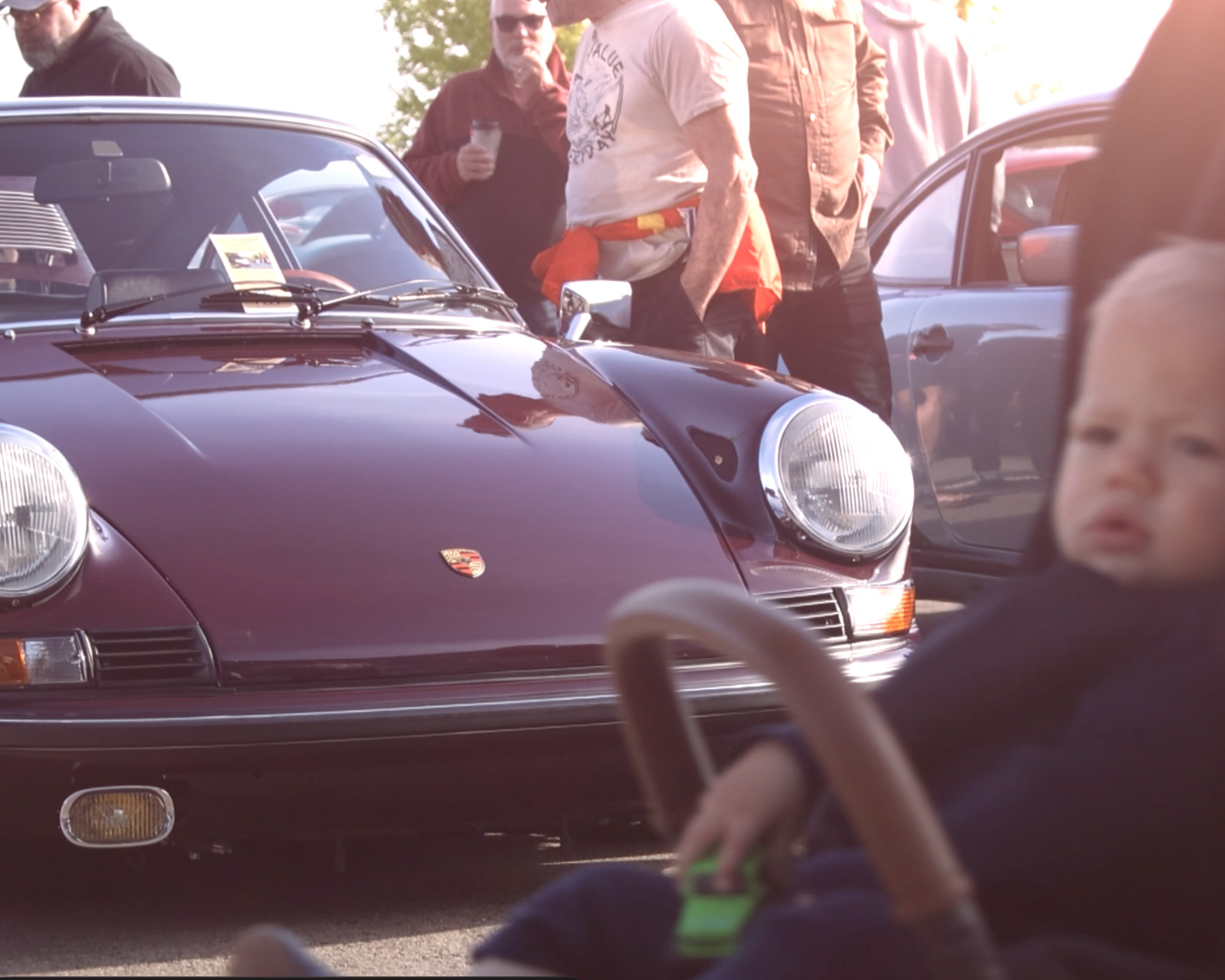 A maroon classic Porsche 911 surrounded by attendees, with a child in a stroller passing in the foreground