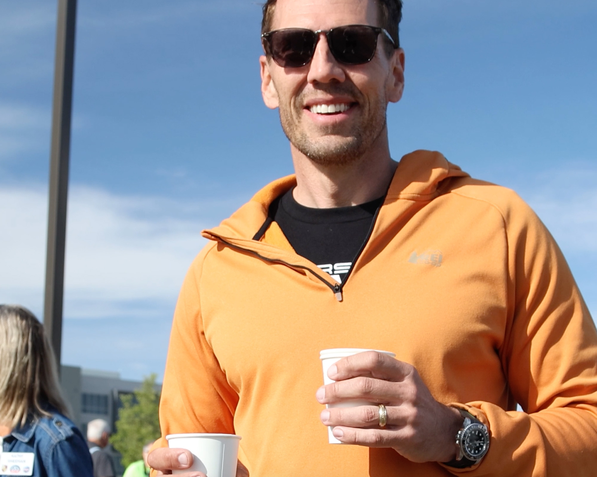 A smiling attendee in an orange hoodie holding two cups of coffee