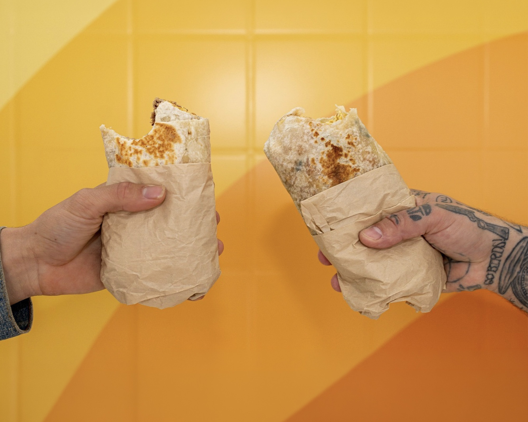 Two Earlybird breakfast burritos wrapped in paper, held up against a yellow wall