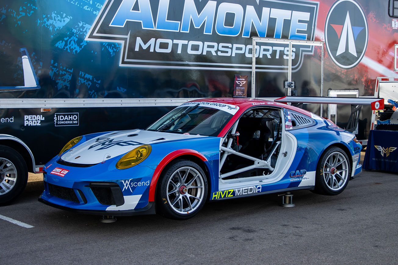 Blue, white and red Porsche 911 race car on jack stands in front of the Almonte Motorsports team trailer at Idaho Air 2026