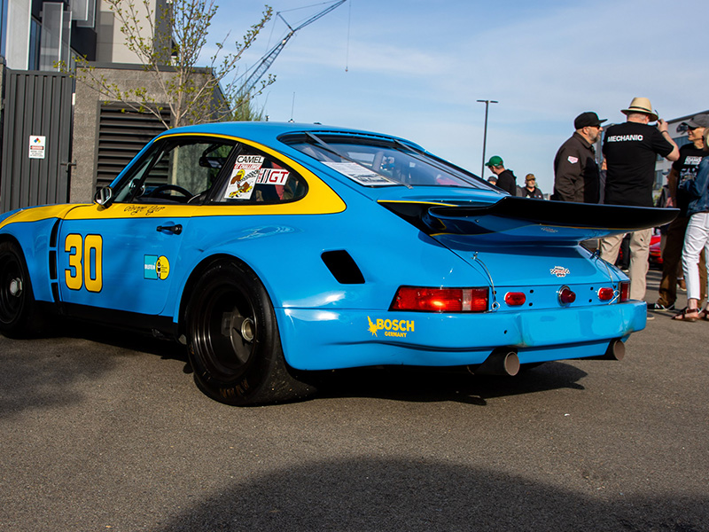 A blue and yellow #30 Porsche 911 race car with a whale tail, parked as spectators chat behind it